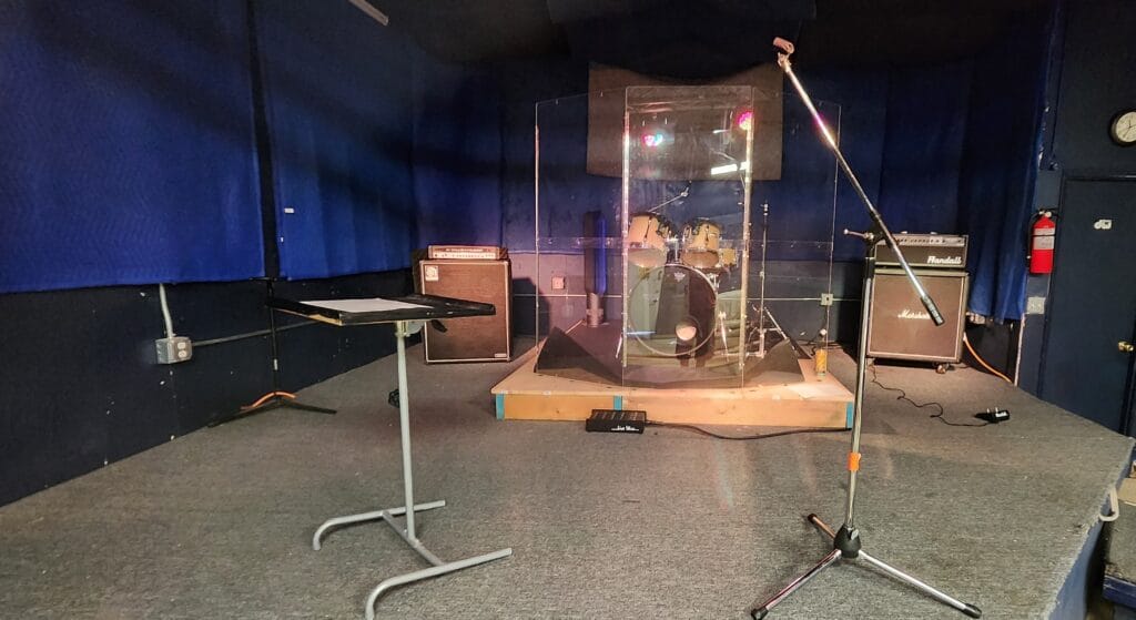 Raised rehearsal stage with drum kit, drum shield, amps, and microphones